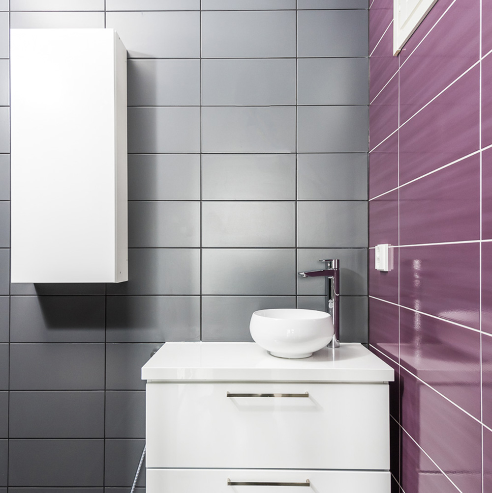 Kylpyhuoneessa on violetit ja harmaat laatat seinässä sekä valkoiset kalusteet.
