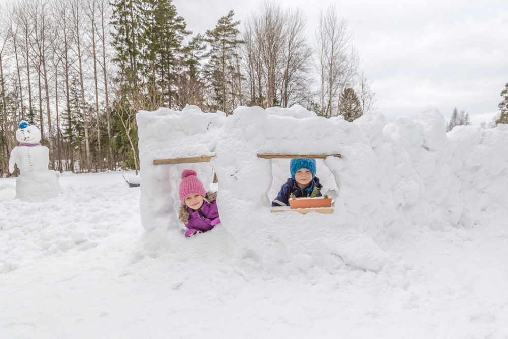 Omakotitalon rakentaminen - Lapset ovar rakentaneet pihalle lumiukkon ja -lumilinnan. He kurkistavat lumilinnan ikkunasta.
