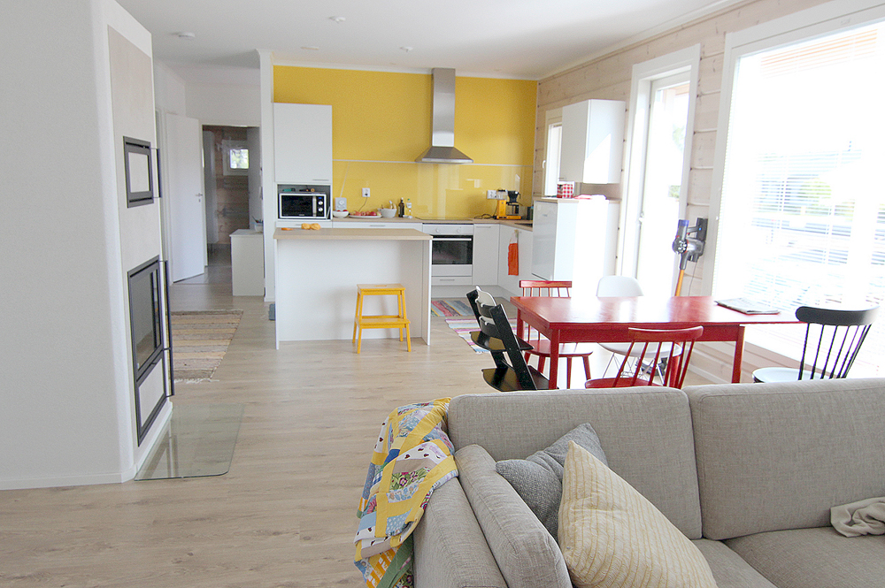 Muuttovalmis hirsitalo: näkymä olohuoneesta keittiöön. Punaisen keittiön pöydän ympärillä on värikkäitä tuolia. Keittiön takaseinä on keltainen.