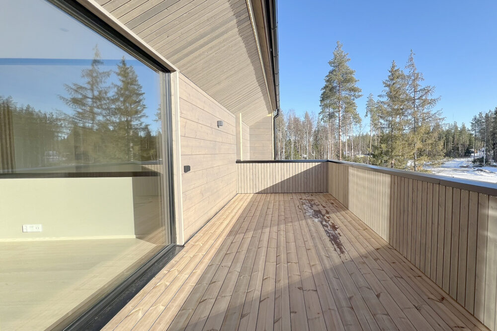 Kaksikerroksinen hirsitalo, tilava parveke kylpee auringon paisteessa.